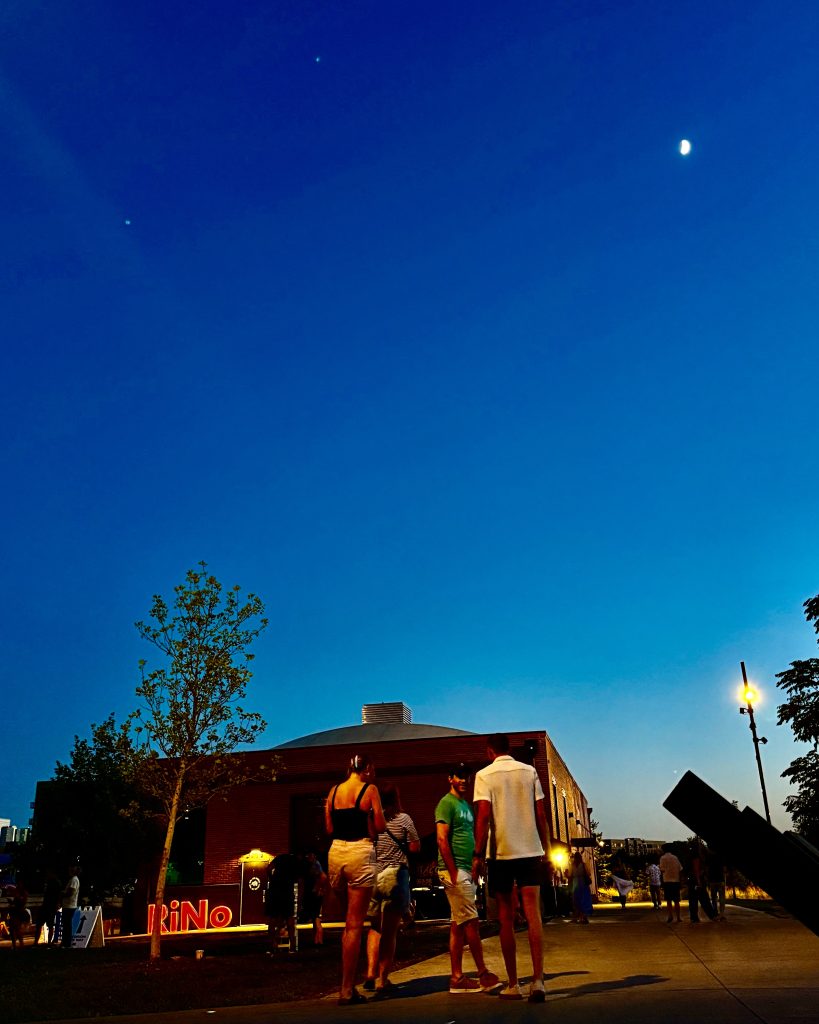 Evening crowd at the RiNo Art Park in Denver under a dusk sky with a crescent moon